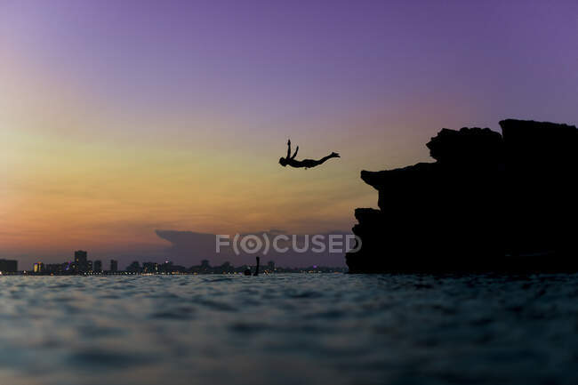 Vista laterale della silhouette della persona che si tuffa nell'acqua dell'oceano dalla pietra — Foto stock
