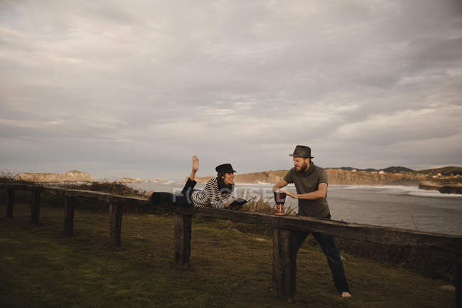 Jeune homme en chapeau jouant sur le tambour main près de femme élégante en chapeau avec bloc-notes et crayon couché sur le siège près de la côte de la mer — Photo de stock