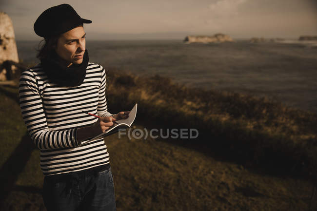 Elegant woman in cap taking notes in notepad on seat on coast near waving sea — Stock Photo