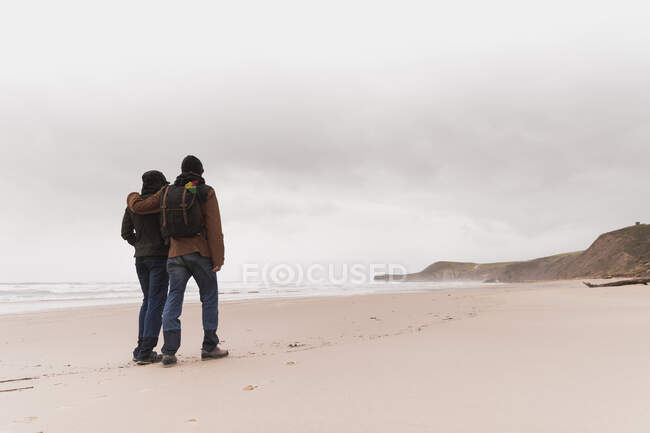 Back view of young couple with backpack in warm wear holding each other on sand beach near sea and hills — Stock Photo