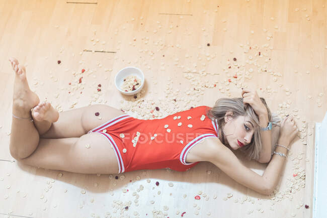 De cima sedutora fêmea descalça olhando para a câmera enquanto deitada no chão perto de muesli derramado — Fotografia de Stock