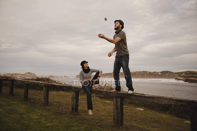 Junger Mann mit Hut jongliert Bälle neben eleganter Frau in Mütze mit Ethiktrommel sitzt auf Sitz in der Nähe der Küste von Meer und bewölktem Himmel — Stockfoto