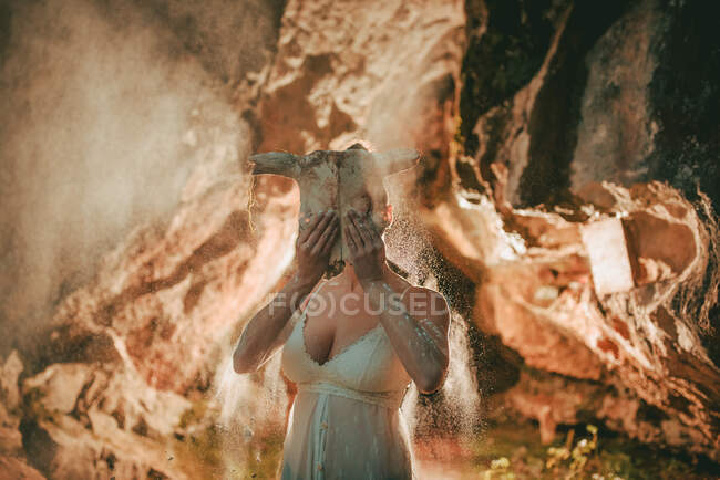 Woman wearing white lace top covering face with animal skull while standing in flying dry dust inside of cave — Stock Photo