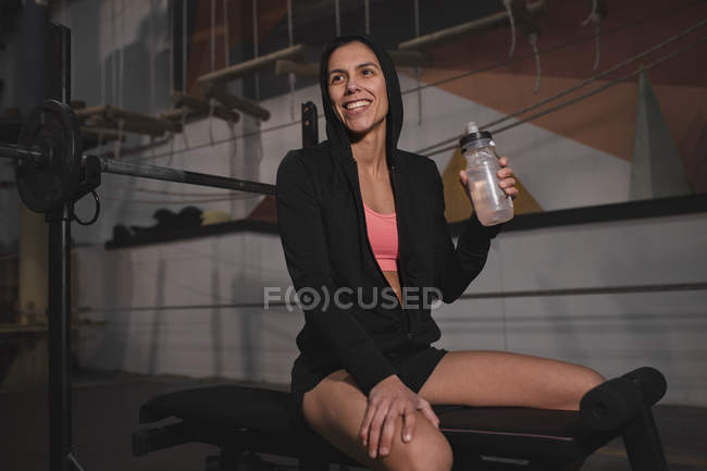 Женщина в спортивной одежде с бутылкой воды сидит на скамейке в тренажерном зале — стоковое фото