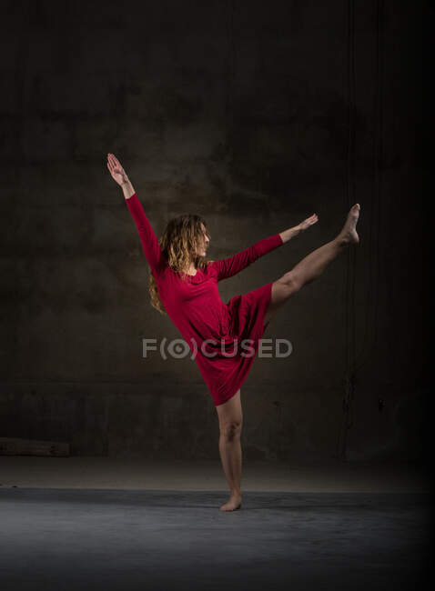 Giovane ballerina magra in abito rosso alzando la gamba e le mani in camera oscura — Foto stock