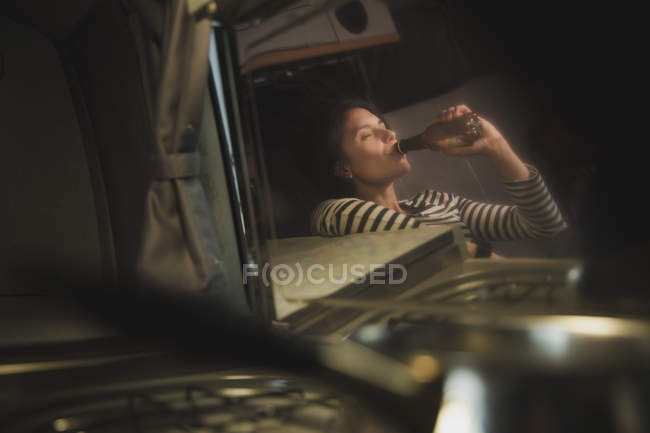 Reflection of young woman with closed eyes drinking from bottle and sitting on sofa near cooker — Stock Photo