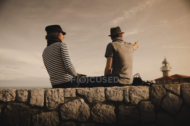 Вид сбоку стильной элегантной пары, сидящей на скалах рядом с маяком и прекрасным небом — стоковое фото