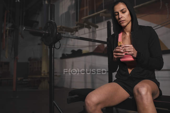 Junge Frau in Sportbekleidung mit Energieriegel sitzt auf Bank im Fitnessstudio — Stockfoto
