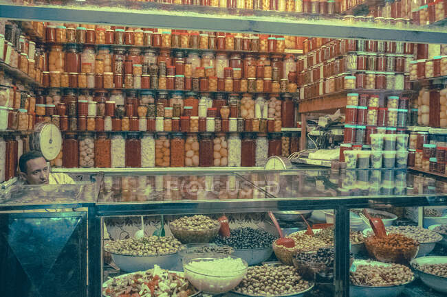31 diciembre 2017 - Marrakech, Marruecos: Hombre adulto sentado en el mostrador con surtido de verduras en escabeche en el mercado - foto de stock
