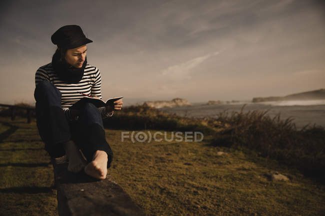 Femme élégante en bonnet prendre des notes dans le bloc-notes et assis sur le siège sur la côte près de la mer ondulante — Photo de stock