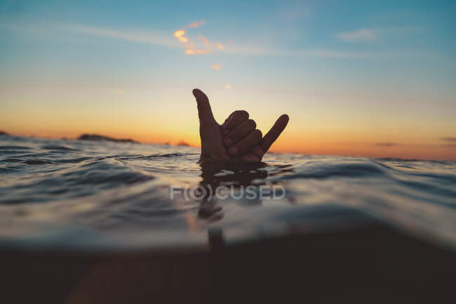 Рука людини, що показує знак шака над поверхнею води з хвилею і блакитним небом увечері на Балі, Індонезія — стокове фото