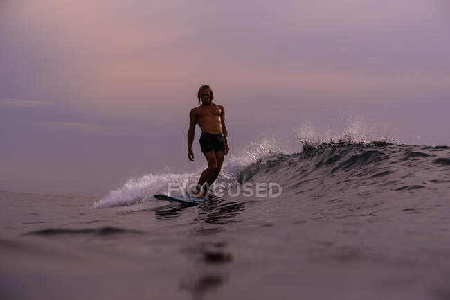 Hombre surfeando entre olas de agua de mar con salpicaduras y cielo nublado por la noche en Bali, Indonesia - foto de stock