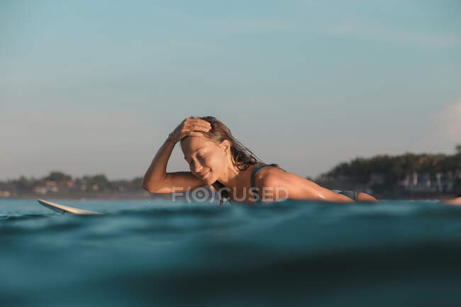 Vista lateral da fêmea alegre flutuando na prancha de surf entre a água do mar e o céu azul em Bali, Indonésia — Fotografia de Stock