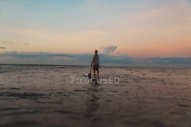 Back view of silhouette of male with paddle on surf board between water of sea and sky in evening on Bali, Indonesia — Stock Photo