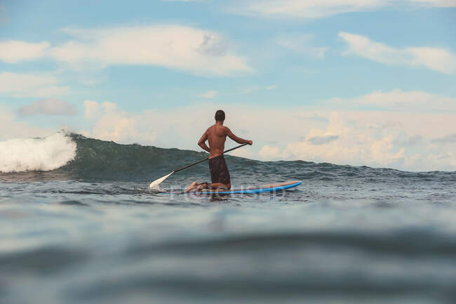 Vista posteriore della pagaia maschile su tavola da surf tra acqua di mare e cielo blu a Bali, Indonesia — Foto stock