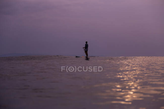 Vista laterale di sagoma maschile con pagaia su tavola da surf tra acqua di mare e cielo in serata a Bali, Indonesia — Foto stock