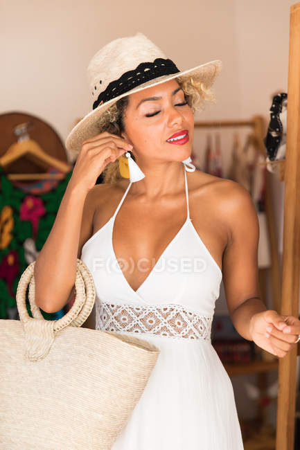 Elegante mujer joven negro con sombrero y bolso de mano que sostiene el pendiente en boutique - foto de stock