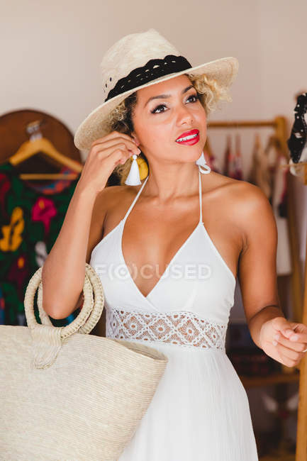 Elegante mujer joven negro con sombrero y bolso de mano que sostiene el pendiente en boutique - foto de stock