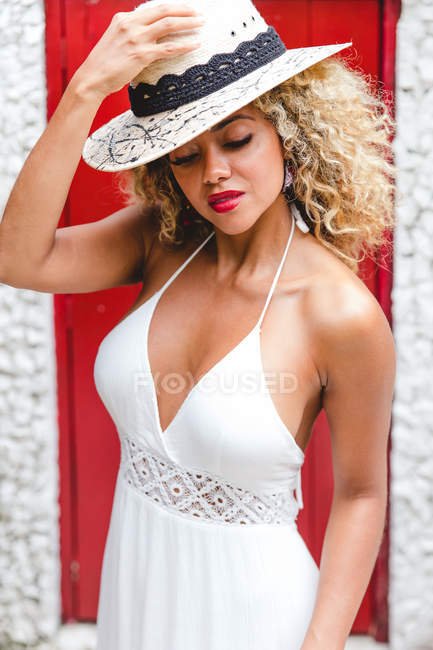 Stylish black young woman with curly hair putting hat on head near wall with red door — Stock Photo