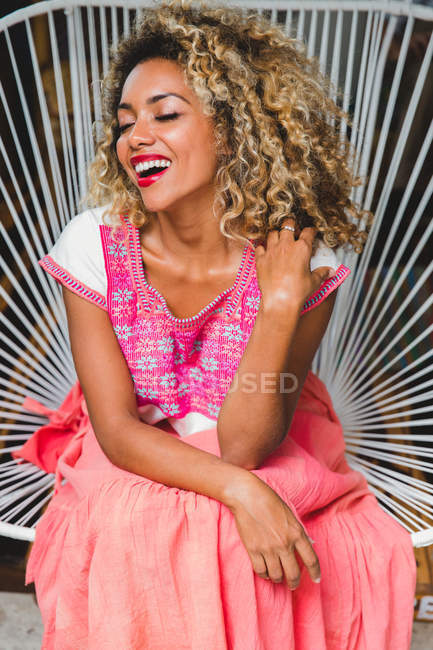 Portrait de jeune femme noire heureuse avec les yeux fermés et les cheveux bouclés assis sur une chaise en osier — Photo de stock
