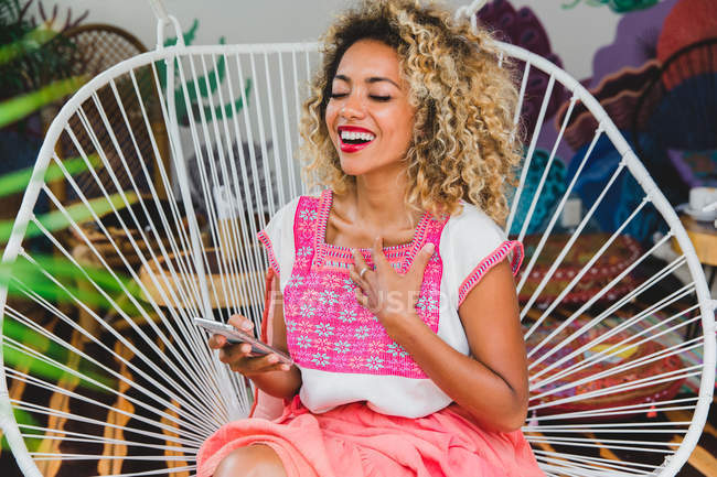 Веселая черная молодая женщина с кудрявыми волосами держит мобильный телефон и сидит на плетеном стуле — стоковое фото