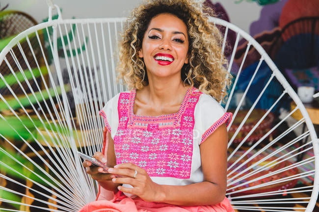 Retrato de una joven negra feliz con el pelo rizado sosteniendo el teléfono móvil y sentada en una silla de mimbre - foto de stock
