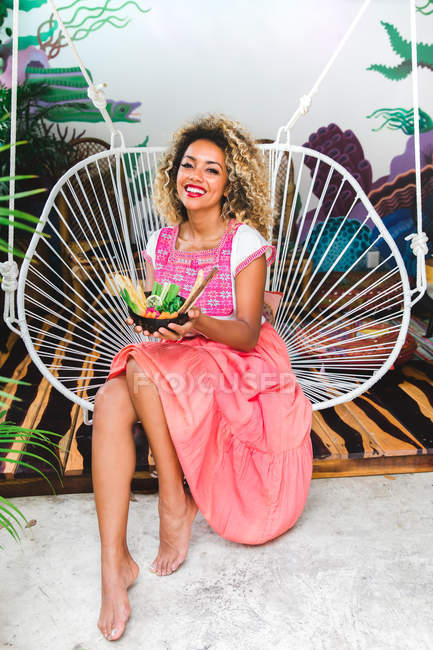 Porträt einer schwarzen jungen Frau, die eine Schüssel mit frischem Gemüse in der Hand hält und auf einem Korbstuhl sitzt — Stockfoto