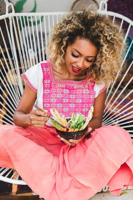 Porträt einer schwarzen jungen Frau, die eine Schüssel mit frischem Gemüse in der Hand hält und auf einem Korbstuhl sitzt — Stockfoto