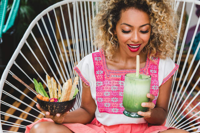 Чорношкіра молода жінка тримає миску зі свіжих овочів, п'є струснути зі скла і сидить на плетеному стільці — стокове фото