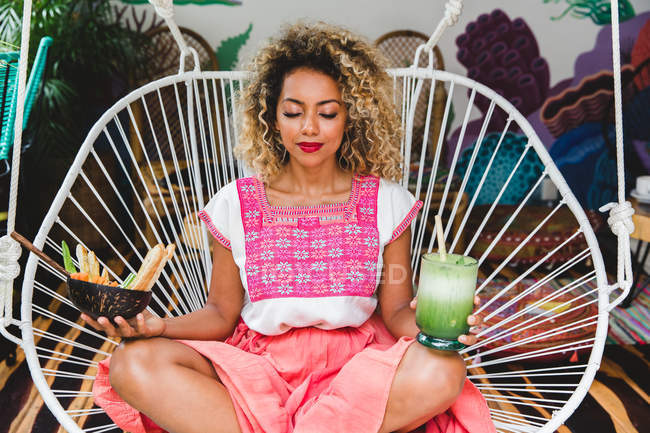 Расслабленная черная молодая женщина держит миску свежих овощей и трясет сидя на плетеном стуле с закрытыми глазами — стоковое фото
