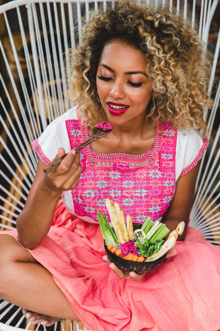 Black young woman eating fresh vegetables with dip in bowl while sitting on wicker chair — Stock Photo