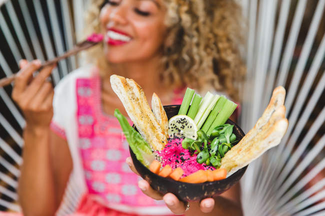 Schwarze junge Frau isst frisches Gemüse mit Dip in Schüssel, während sie auf einem Korbstuhl sitzt — Stockfoto