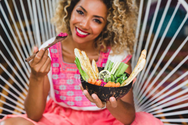 Черная молодая женщина ест свежие овощи с соусом в миске, сидя на плетеном стуле — стоковое фото