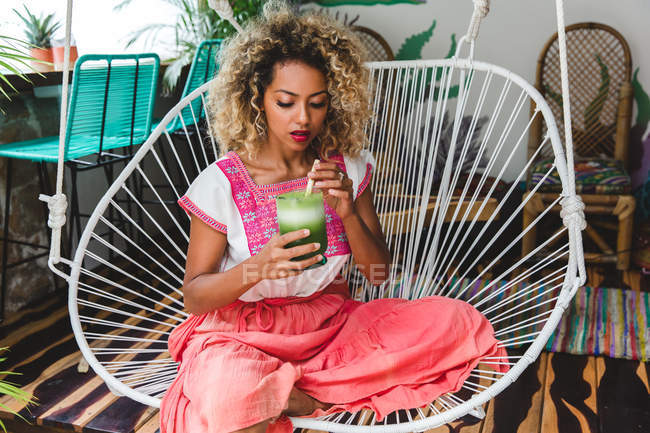 Чорношкіра молода жінка тримає склянку струсу, сидячи на плетеному стільці — стокове фото