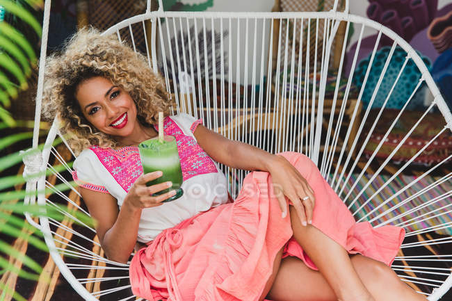 Portrait de jeune femme noire heureuse tenant un verre de shake sur une chaise en osier — Photo de stock