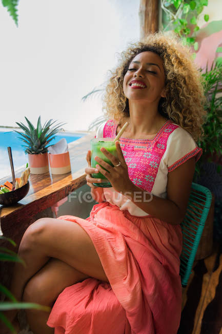 Happy black young woman holding glass of shake and sitting near wooden table with bowl of vegetables in cafe — Stock Photo