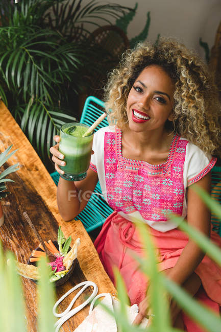 Felice giovane donna nera in possesso di un bicchiere di scossa e seduta vicino a un tavolo di legno con ciotola di verdure nel caffè — Foto stock