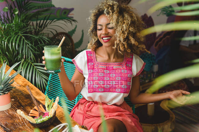 Веселая черная молодая женщина держит стакан коктейля и сидит возле деревянного стола с миской овощей в кафе — стоковое фото