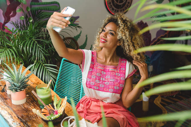Souriant jeune femme noire avec verre de shake et bol de salade prendre selfie avec téléphone portable dans le café — Photo de stock
