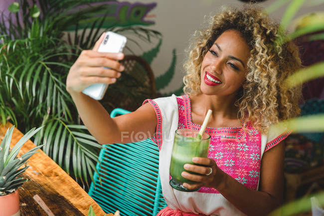 Улыбающаяся черная молодая женщина со стаканом коктейля делает селфи с мобильным телефоном в кафе — стоковое фото