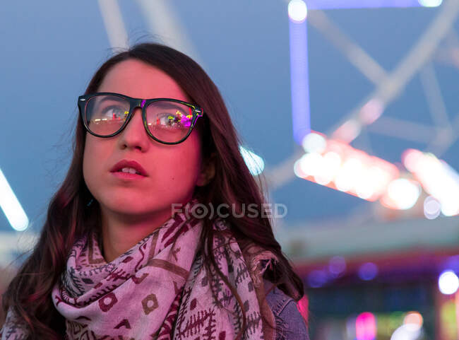 Allegro signora in fascia e occhiali vicino evidenziato ruota panoramica nel parco divertimenti in serata su sfondo sfocato — Foto stock
