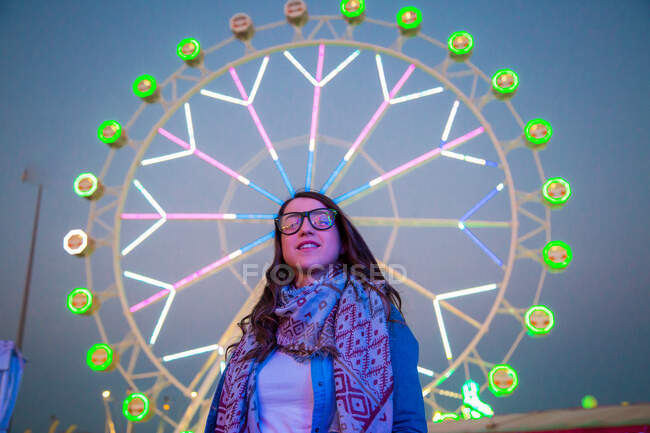 Cheerful lady in sash and eyeglasses near highlighted ferris wheel in amusement park in evening on blurred background — Stock Photo