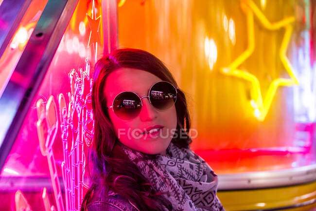Модна впевнена дама в раковинах і сонцезахисних окулярах біля стіни з неоновими вогнями на вулиці вночі — стокове фото
