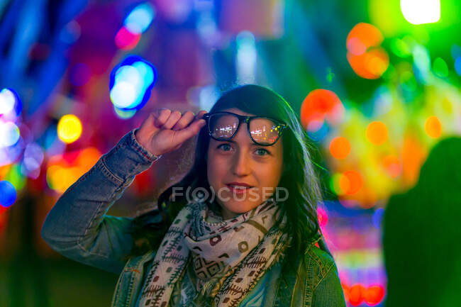 Senhora confiante na moda em faixa e óculos de sol perto de luzes de néon na rua à noite no fundo borrado — Fotografia de Stock