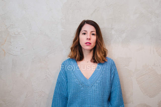 Lovely serious young woman in knitted sweater looking at camera and standing near grey wall — Stock Photo