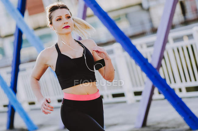 Mujer en ropa deportiva con auriculares y smartphone corriendo en la calle - foto de stock