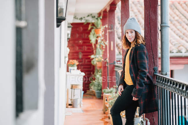 Mujer joven atractiva positiva en ropa abrigada y sombrero mirando hacia otro lado y de pie cerca de la casa y la cerca - foto de stock