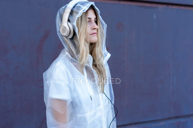 Nachdenkliche junge Frau in Regenmantel, die mit Kopfhörern Musik hört und in der Nähe einer Mauer auf der Straße wegschaut — Stockfoto