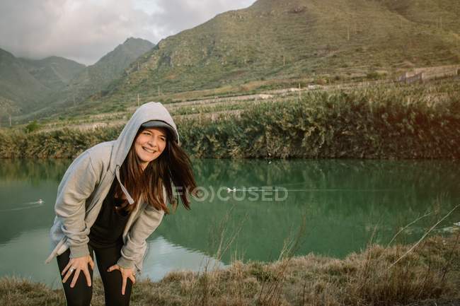 Mujer positiva teniendo un descanso después de correr en la naturaleza cerca del lago y las montañas - foto de stock
