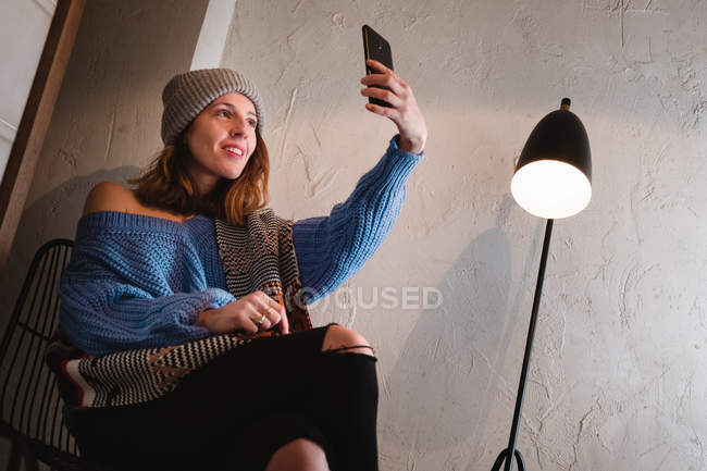 Молодая женщина в вязаном свитере с шарфом и шляпой делает селфи на смартфоне и сидит на стуле возле стены и лампы в комнате — стоковое фото